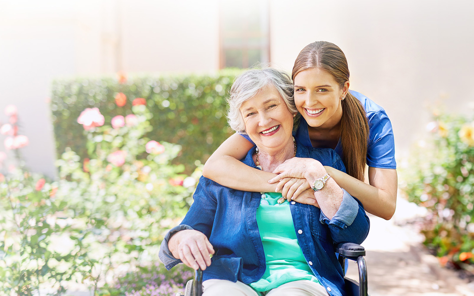 An elderly woman in a wheelchair is smiling and hugging a young adult woman who is standing beside her, both are outdoors with flowers and greenery in the background.