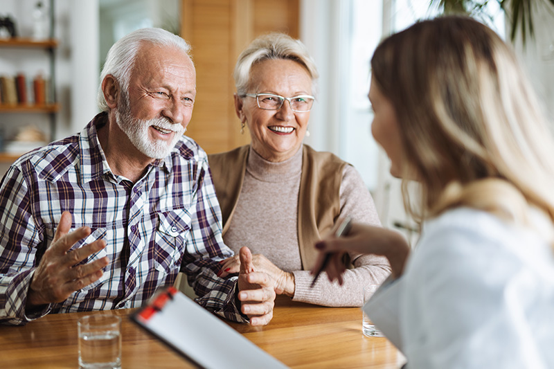 Two elderly people, a man and a woman, seated at a dining table with a laptop in front of them they are engaged in conversation with a young female figure who appears to be a waitress or service provider.